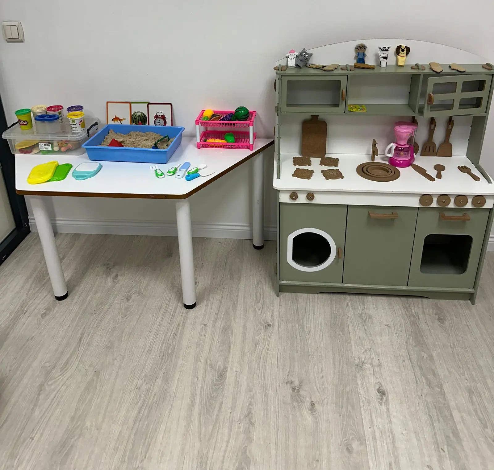 Oyun terapisi odasında oyun hamuru ve kum tepsisi bulunan masa ile ahşap oyuncak mutfak